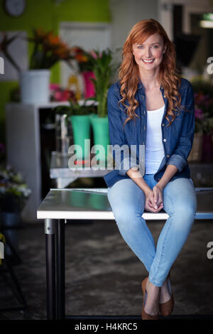 Happy female florist sitting on table in flower shop Banque D'Images