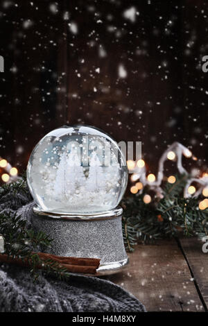 L'image rustique d'un globe de neige entouré par des branches de pins, des bâtons de cannelle et un foulard gris chaud avec des flocons de neige tomber doucement. Banque D'Images