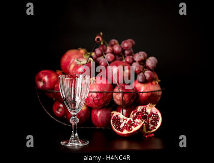 Nature morte sur un fond sombre. (Vin) verres d'alcool, de fruits et de petits fruits (pommes, raisins et grenades) dans le panier. Banque D'Images