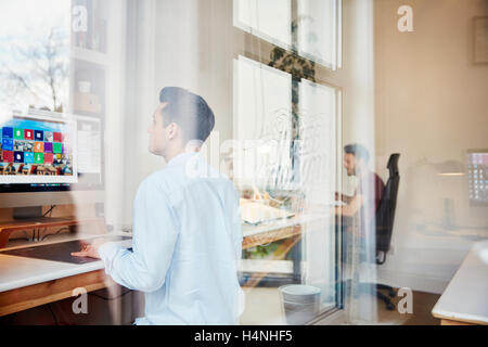 Deux hommes travaillant sur des ordinateurs dans un studio de design, vue à travers une fenêtre et réflexions de la fenêtre et les arbres. Banque D'Images