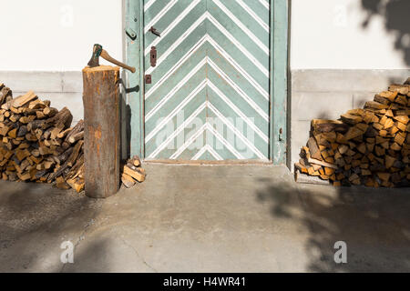 Détail d'un bâtiment rural : une porte en bois verdâtre avec des rayures blanches, bois empilés et un couperet avec une hache. Banque D'Images