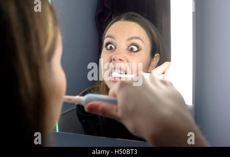 Jeune femme à la recherche dans la salle de bains miroir car elle nettoie les dents avec brosse à dents électrique Banque D'Images