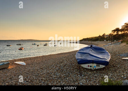 Coucher du soleil à Ringstead Bay, Dorset, UK, voile bateaux amarrés dans la baie et un canot couvert de toile goudronnée sur la plage de galets à l'avant-plan Banque D'Images