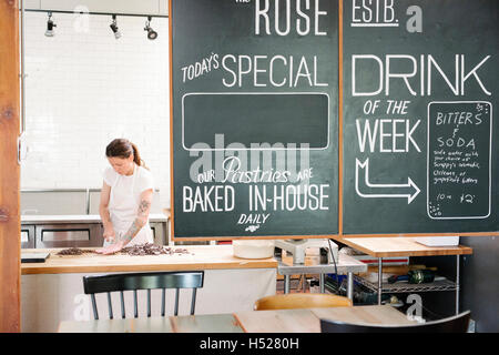 Femme portant un tablier blanc à un comptoir d'une boulangerie, menu sur un tableau noir. Banque D'Images