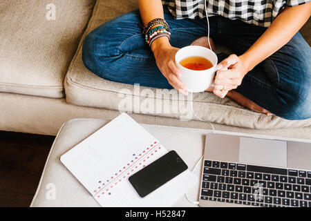 Le port de jeans femme assise sur un canapé en face d'un ordinateur portable, tenant une tasse. Banque D'Images