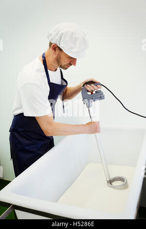 L'homme dans une crémerie, le port de tablier et capuchon de protection, en remuant le lait de chèvre, fromage de chèvre faire. Banque D'Images