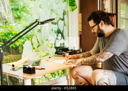 Homme barbu avec des tatouages sur ses bras et jambes assis à un bureau, dessin. Banque D'Images