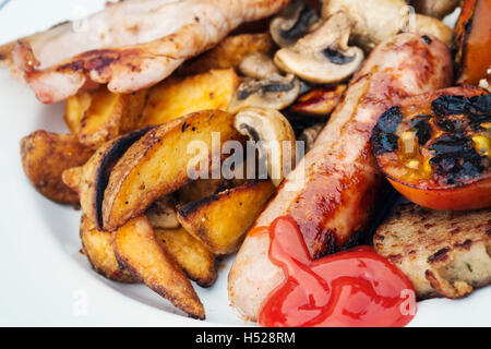 Gros plan d'une assiette de nourriture, petit déjeuner anglais, bacon, champignons, saucisses, tomates, pommes de terre grillées et sauce tomate. Banque D'Images