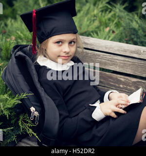 Belle fille blonde dans un uniforme scolaire siège au banc de parc en graduation cap et la lecture de livre. Banque D'Images