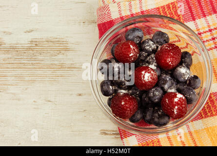 Bleuets et framboises saupoudrées de sucre glace dans un bol en verre sur un tissu coloré et mouvementé, ancienne table en bois clair Banque D'Images