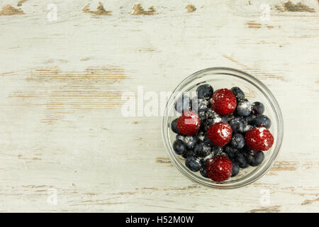 Bleuets et framboises saupoudrées de sucre glace dans un bol en verre sur une vieille table en bois clair, dans un style vintage , spa copie Banque D'Images