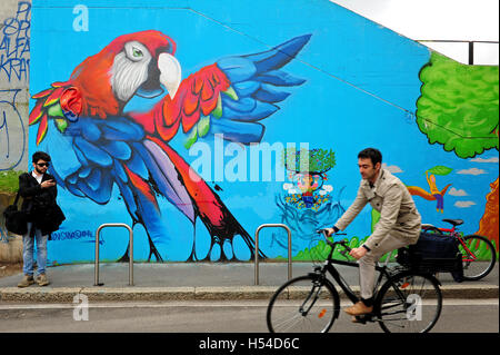 Un homme sur un vélo en face d'une peinture murale géante avec un perroquet, dans le quartier Isola à Milan. Banque D'Images