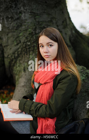 Cute girl assis sur un fond d'un vieil arbre énorme. Un foulard rouge vif attire l'attention. Sur un circuit à la jeune fille l'ouvrir aucun Banque D'Images