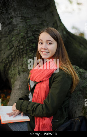 Cute girl assis sur un fond d'un vieil arbre énorme. Un foulard rouge vif attire l'attention. Sur un circuit à la jeune fille l'ouvrir aucun Banque D'Images