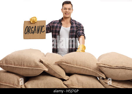 Jeune agriculteur debout derrière une pile de sacs de toile et tenant un panneau qui dit carton organic isolé sur fond blanc Banque D'Images