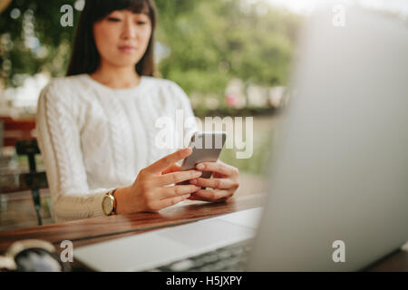 Shot of asian woman using mobile phone at outdoor cafe. Jeune femme assise à table avec coffre de la lecture de texte message sur son marquage ce Banque D'Images