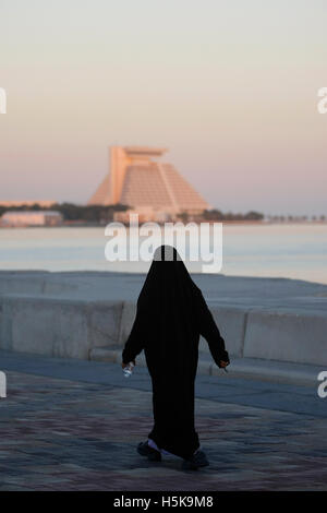 Femme arabe portant une robe noire traditionnelle, marcher le long de la Corniche, l'hôtel Sheraton à l'arrière, District de West Bay, Doha Banque D'Images