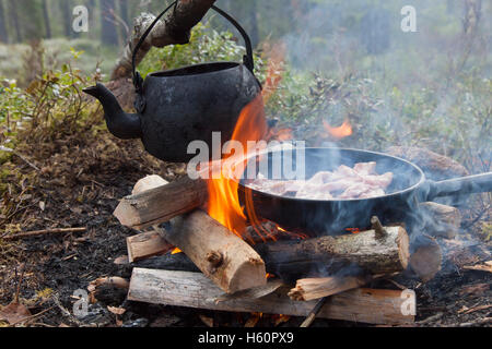 Étain noirci électrique de l'eau bouillante et cuire le bacon sur pan flammes de feu de camp au cours de la randonnée en forêt Banque D'Images