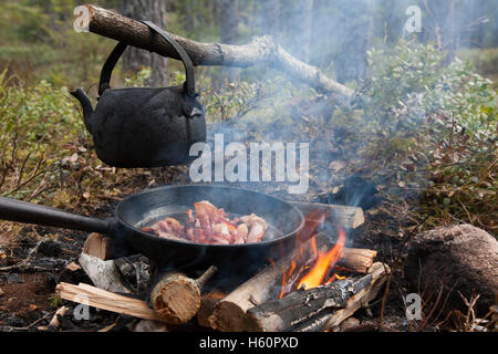 Étain noirci électrique de l'eau bouillante et cuire le bacon sur pan flammes de feu de camp au cours de la randonnée en forêt Banque D'Images