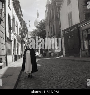 Années 1950, historiques, une religieuse catholique promenades dans un quartier calme de la rue pavées, avec les dômes de la célèbre cathédrale française, Sacre-Coeur, dans l'arrière-plan, Montmartre, Paris, France. Banque D'Images