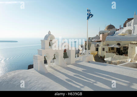 Ombre de cloches sur un toit de l'église à Oia, Santorin avec un drapeau grec battant contre un ciel bleu. Banque D'Images