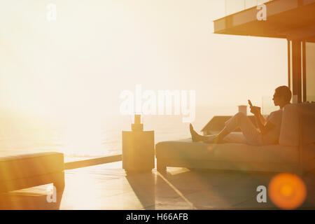 Silhouette woman using cell phone sur une chaise longue sur le luxe d''un balcon avec vue sur l'océan au coucher du soleil Banque D'Images