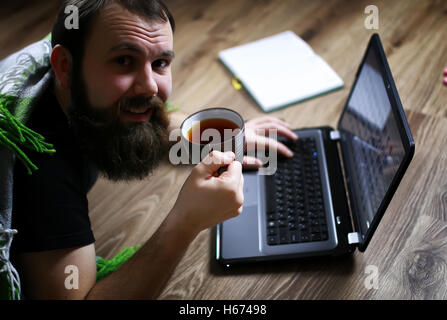 Bel homme barbu se trouve sur le sol enveloppée dans un plaid, ordinateur portable et de travail Banque D'Images