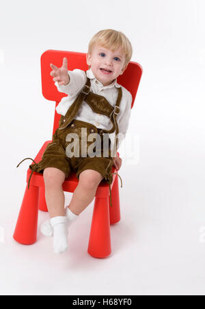 Little Boy wearing Lederhose bavarois traditionnel, short en cuir, assis sur une chaise rouge Banque D'Images