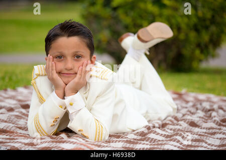 Jeune garçon avec White Sailor Costume dans sa Première Communion allongé sur une couverture sur l'herbe reposant sa tête sur ses mains et regarder Banque D'Images