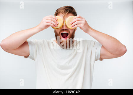 Homme barbu excité couvrant les yeux avec des hamburgers isolé sur fond blanc Banque D'Images