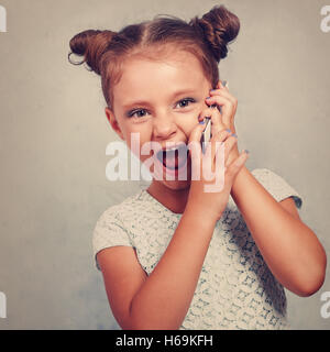 Heureux kid girl talking on mobile phone avec bouche ouverte sur fond bleu. Tons Vintage closeup portrait Banque D'Images