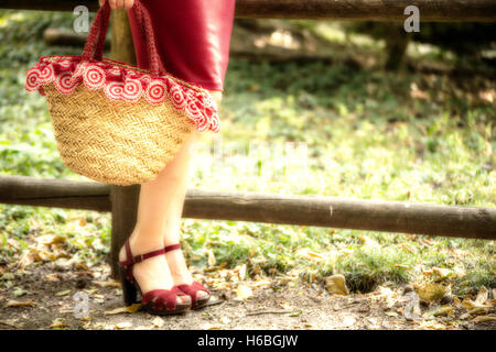 Et bien longue jambes féminine d'une femme qui est en attente dans un parc, elle porte une robe tube rouge, sandales à talons hauts et tenant un sac rempli de fleurs dans un style campagnard Banque D'Images
