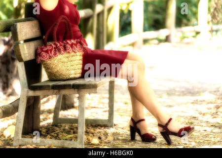 Et bien longue jambes féminine d'une femme qui attend sur un banc dans un parc, elle porte une robe tube rouge, sandales à talons hauts et tenant un sac rempli de fleurs dans un style campagnard Banque D'Images