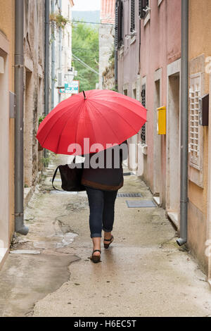 Une femme portant un parapluie rouge vif dans une petite ruelle de Ston en Croatie sous un ciel couvert et journée de bruine Banque D'Images