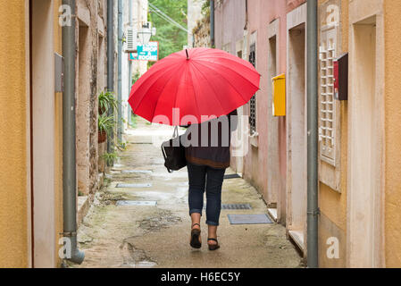 Une femme portant un parapluie rouge vif dans une petite ruelle de Ston en Croatie sous un ciel couvert et journée de bruine Banque D'Images