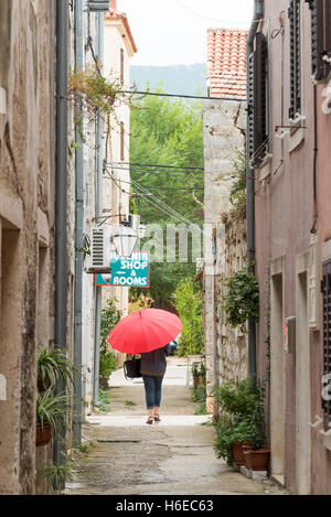 Une femme portant un parapluie rouge vif dans une petite ruelle de Ston en Croatie sous un ciel couvert et journée de bruine Banque D'Images