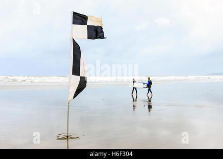 Deux surfers carrying une planche de surf à la mer à marée basse sur la plage à Westward Ho !, Devon, England, UK, un drapeau noir et blanc à l'avant-plan Banque D'Images