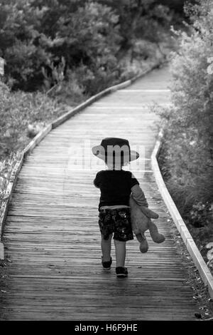 Deux ans boy wearing hat exerçant son ours marche sur la promenade à travers les dunes de sable avec des arbres de chaque côté , Australie Banque D'Images