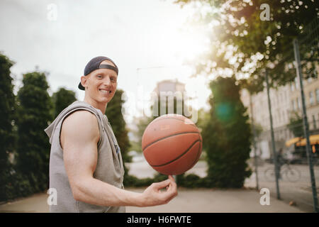 Portrait of smiling streetball player tourner la balle sur une cour. Heureux jeune homme basket-ball d'équilibrage sur son doigt. Banque D'Images