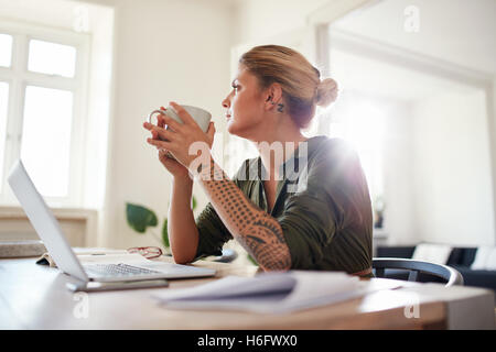 Tourné à l'intérieur de la jeune femme avec du café à l'écart et la pensée. Femme sérieuse assis à votre bureau à domicile. Banque D'Images