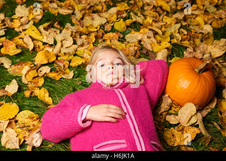 Enfant mignon fille blonde allongée dans les feuilles d'automne à la citrouille. Banque D'Images