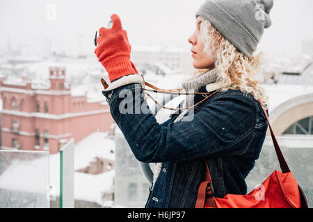 Jeune femme blonde curly dans des vêtements chauds, des gants et des sacs rouge debout sur le toit et le tir sur caméra film retro Banque D'Images