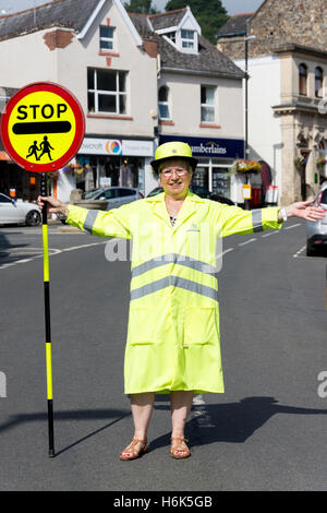 Lollipop lady (agent de patrouille de passage de l'école) l'arrêt de la circulation sur la rue de l'avancement, Bovey Tracey, Devon, Angleterre, Royaume-Uni Banque D'Images