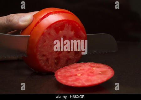 Close Up de trancher une grande personne, mûres, tomates biologiques avec un couteau dentelé sur un comptoir brun foncé Banque D'Images