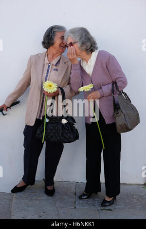 Deux personnes âgées portugais chers vêtus de vêtements semblables holding Flowers et gros sacs à main le partage d'un secret. Banque D'Images