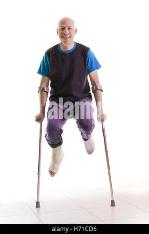 Jeune homme avec une cheville cassée et un livre blanc sur la jambe en sautant et en s'amusant avec ses béquilles (isolé sur blanc) Banque D'Images