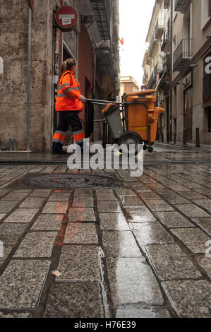 Femme balai avec des vêtements de travail orange, marche avec un chariot de nettoyage municipal, à travers les rues de Valence, Espagne, avec le plancher pavé humide Banque D'Images