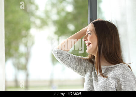 Vue latérale d'un happy girl laughing with hand on head et à l'extérieur à travers une fenêtre à la maison ou chambre d'hôtel Banque D'Images