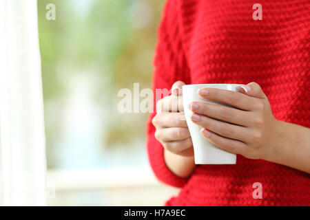 Close up of a woman wearing red pullover avec mains tenant une tasse de café à côté d'une fenêtre avec un fond vert à l'extérieur Banque D'Images