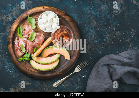 Prosciutto di Parma, cantaloup, melon mozzarella dans un bol et verre de vin rose en plateau de service rond sur bleu foncé plis Banque D'Images